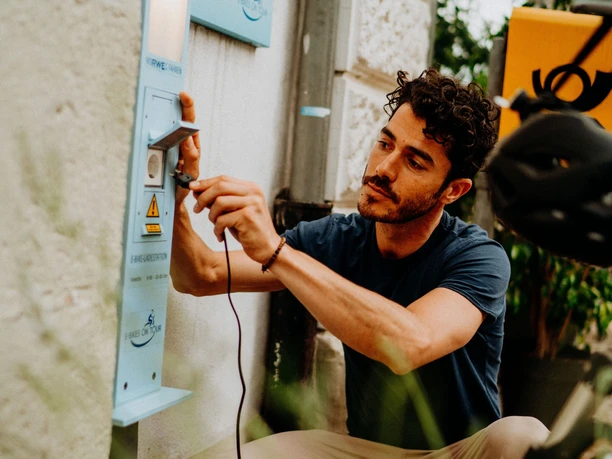 E-Bike Aufladestation Ein Mann lädt sein E-Bike an einer öffentlichen Ladestation im Freien auf, umgeben von Pflanzen.