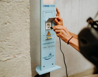 E-Bike Aufladestation Eine Person schließt ein Ladekabel an eine E-Bike-Ladestation an einer hellen Wand an.