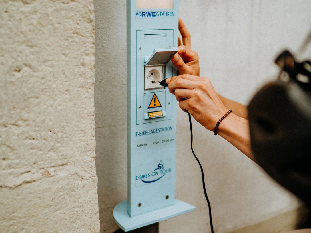 E-Bike Aufladestation Eine Person schließt ein Ladekabel an eine E-Bike-Ladestation an einer hellen Wand an.