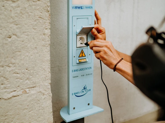E-Bike Aufladestation Eine Person schließt ein Ladekabel an eine E-Bike-Ladestation an einer hellen Wand an.