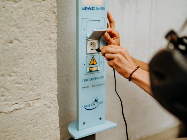 E-Bike Aufladestation Eine Person schließt ein Ladekabel an eine E-Bike-Ladestation an einer hellen Wand an.
