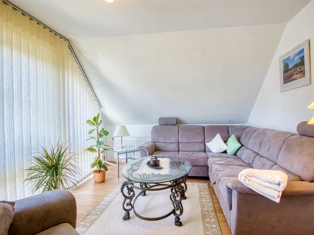 Sitzecke-pallme.jpeg Helles Wohnzimmer mit Dachschräge, braunem Ecksofa, großem Fenster und Glastisch.