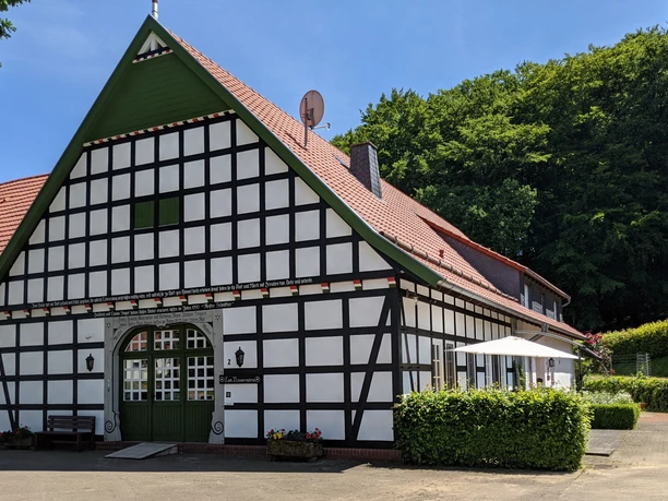 Ein traditionelles Fachwerkhaus mit rotem Dach in ländlicher Umgebung, umgeben von grüner Natur.