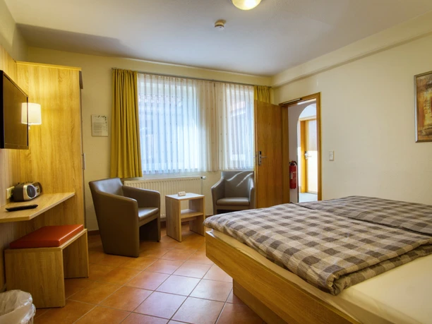 Helles Doppelzimmer mit Fliesenboden, Holzbett, zwei Sesseln am Fenster und TV.