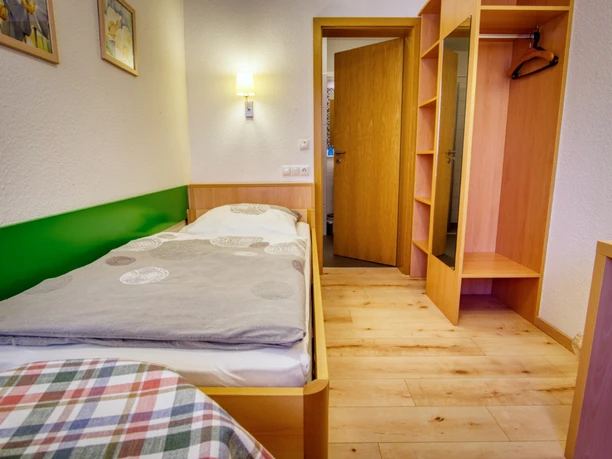 Einzelzimmer-thieshof (2).jpg Helles Einzelzimmer mit Holzboden, Bett, offenem Holzschrank mit Spiegel und Zugang zum Bad.