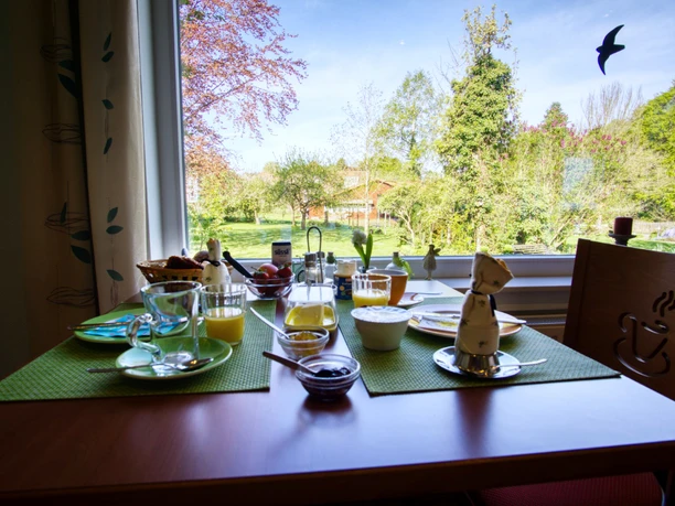 Gedeckter Frühstückstisch am Fenster mit Blick in einen sonnigen, grünen Garten.