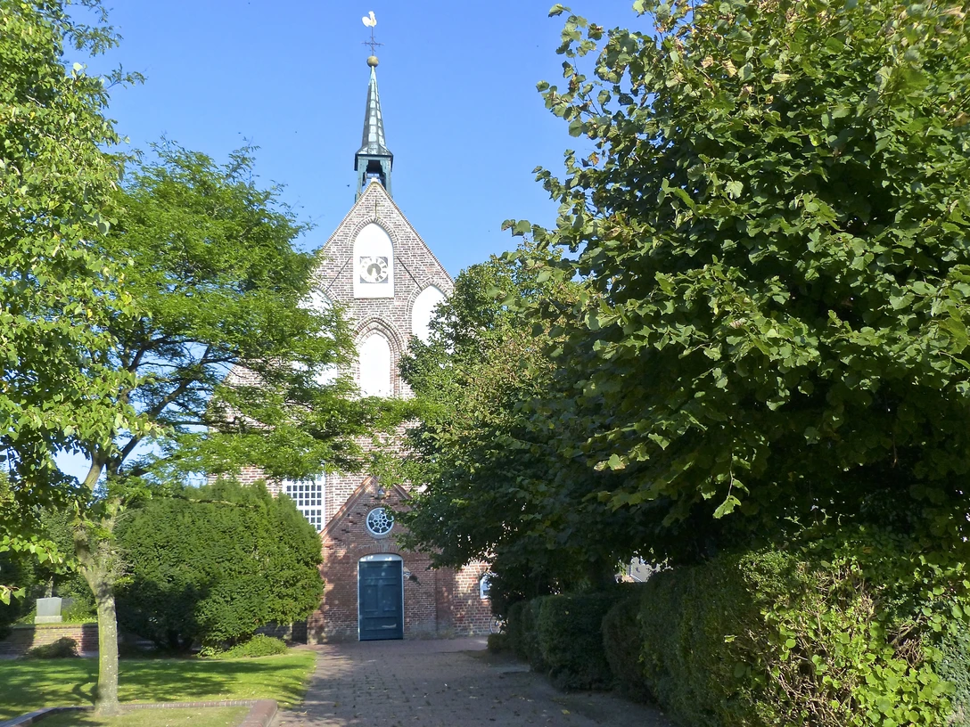 Manslagt Backsteinkirche in Manslagt mit grünem Eingangstor, umgeben von Bäumen unter blauem Himmel.