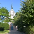 Manslagt Backsteinkirche in Manslagt mit grünem Eingangstor, umgeben von Bäumen unter blauem Himmel.