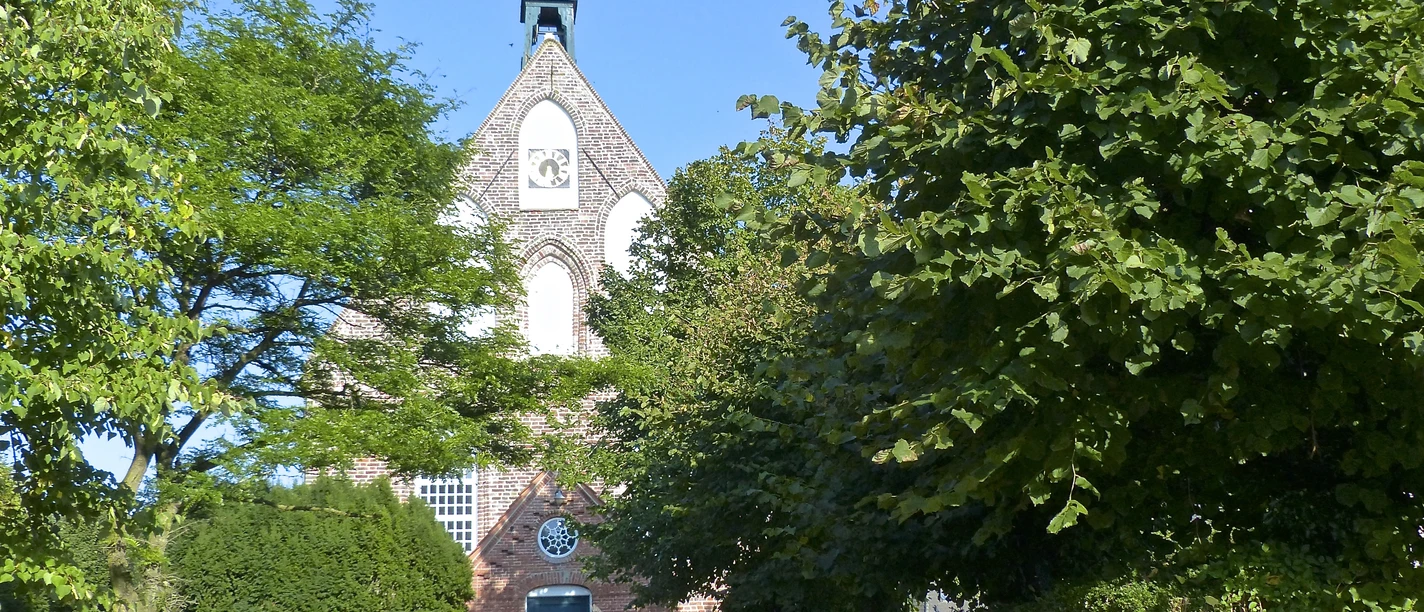 Manslagt Backsteinkirche in Manslagt mit grünem Eingangstor, umgeben von Bäumen unter blauem Himmel.