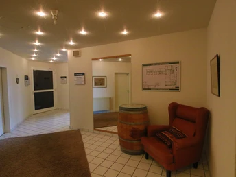 Ein Flur mit weißen Wänden, einem Fass als Tisch, einem roten Sessel und Deckenspots beleuchtet.