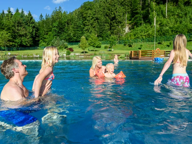 Alpenschwimmbad Oberau Nichtschwimmerbecken Familie badet im flachen Becken vor grünem Hang und Holzsteg an einem sonnigen Tag.