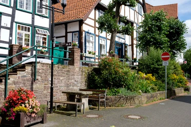Fachwerkhäuser mit roten Ziegeldächern und bunter Blumenschmuck prägen eine ruhige Straßenszene.