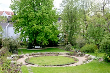 Kreisförmige Rasenfläche im Garten, umgeben von Bäumen und Sträuchern mit einer Steinmauer.