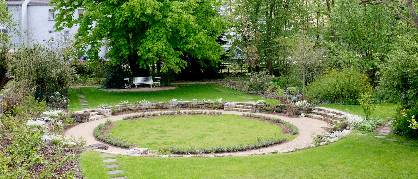 Kreisförmige Rasenfläche im Garten, umgeben von Bäumen und Sträuchern mit einer Steinmauer.