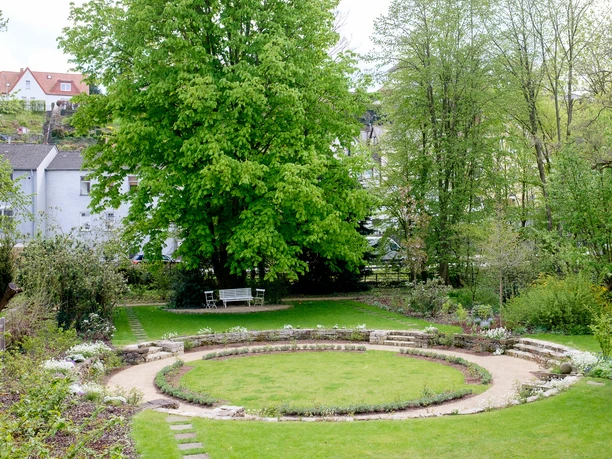 Garten Malz Kreisförmige Rasenfläche im Garten, umgeben von Bäumen und Sträuchern mit einer Steinmauer.