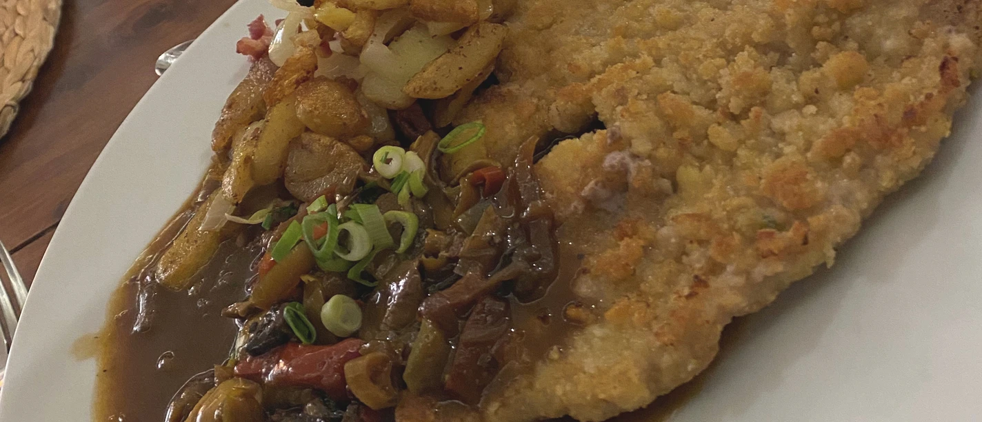 A golden fried Jägerschnitzel on a white plate with mushroom sauce and fried potatoes.