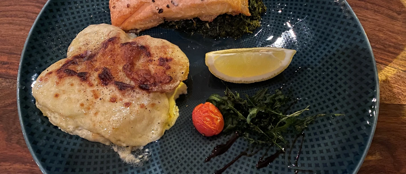 Lachsgericht Lachsfilet auf einem Bett aus grünem Gemüse mit Kartoffelgratin, Kirschtomate und Zitronenspalte.