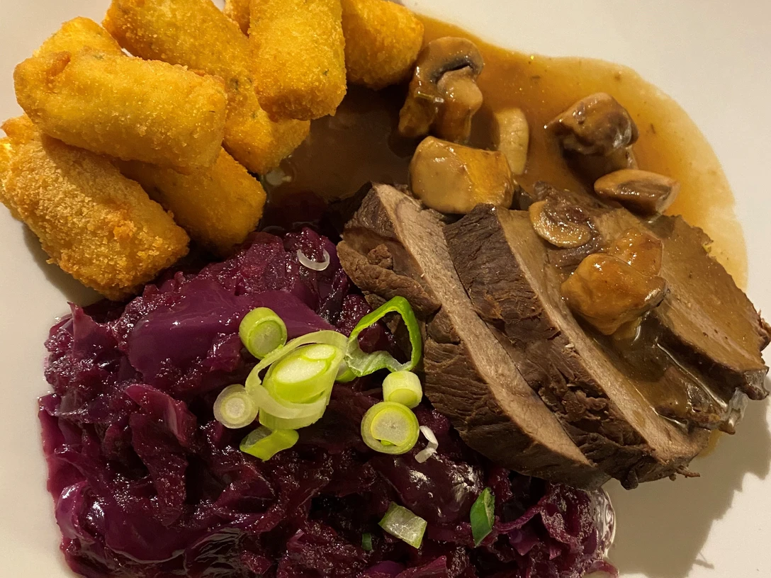 Hirschbraten Ein Teller mit Hirschbraten, Rotkohl, Kroketten und Pilzsoße, garniert mit Frühlingszwiebeln.