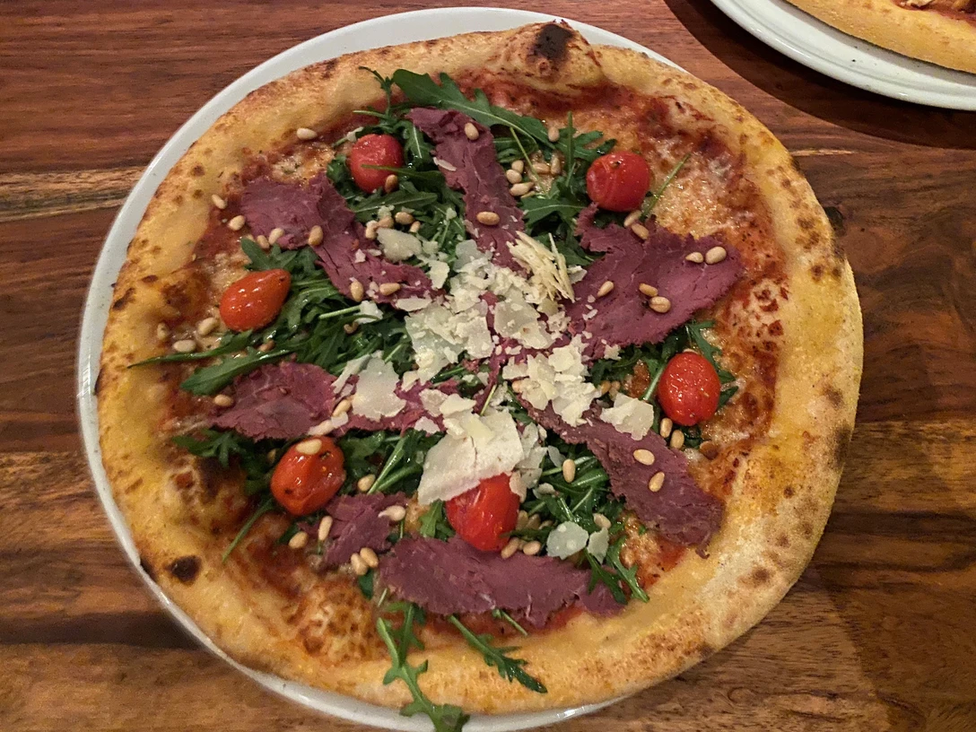 Pizza belegt mit dünnen Fleischscheiben, Rucola, Kirschtomaten, Pinienkernen und Parmesanspänen.Pizza topped with thin slices of meat, rocket, cherry tomatoes, pine nuts and Parmesan shavings.Pizza toppet med tynde skiver kød, rucola, cherrytomater, pinjekerner og parmesanspåner.Pizza belegd met dunne plakjes vlees, rucola, kerstomaatjes, pijnboompitten en Parmezaanschaafsel.
