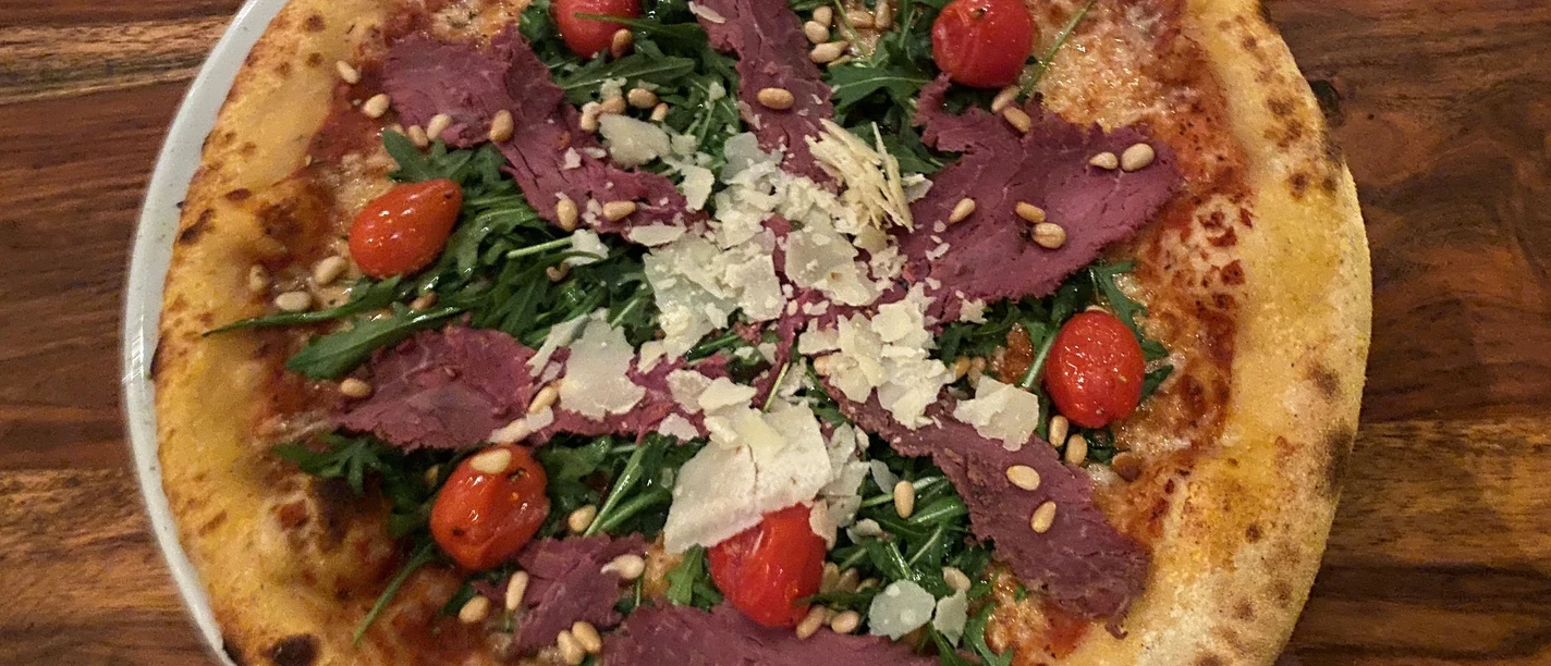 Pizza Pizza belegt mit dünnen Fleischscheiben, Rucola, Kirschtomaten, Pinienkernen und Parmesanspänen.