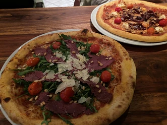 Pizza Zwei Pizza auf Holztisch, vorn mit Rucola, Kirschtomaten, Parmesanspänen und Pinienkernen garniert.