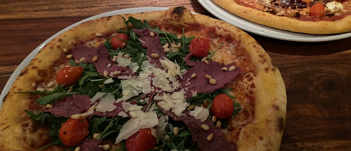 Pizza Zwei Pizza auf Holztisch, vorn mit Rucola, Kirschtomaten, Parmesanspänen und Pinienkernen garniert.