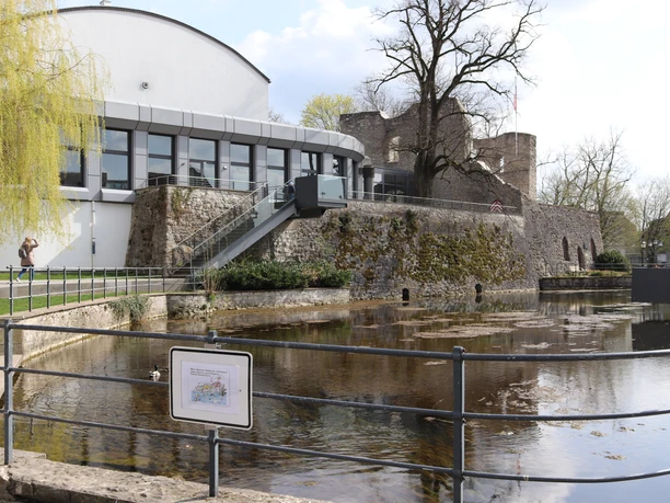 IMG_0124.JPG Historische Stadtmauer mit Burggraben und modernem Gebäude, umgeben von Bäumen und Wasserfläche.