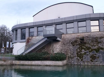 Lippe1.jpg Modernes Café-Restaurant mit grauer Fassade auf einem Steinbau an einem ruhigen, klaren Gewässer.