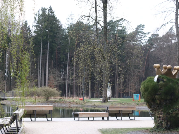 IMG_0619.JPG Ein ruhiger Park mit Bänken am Ufer eines kleinen Sees, umgeben von hohen Bäumen und Skulpturen.