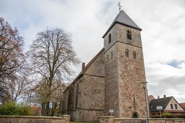 A historic sandstone church with a tower and a pointed roof, surrounded by trees and houses.