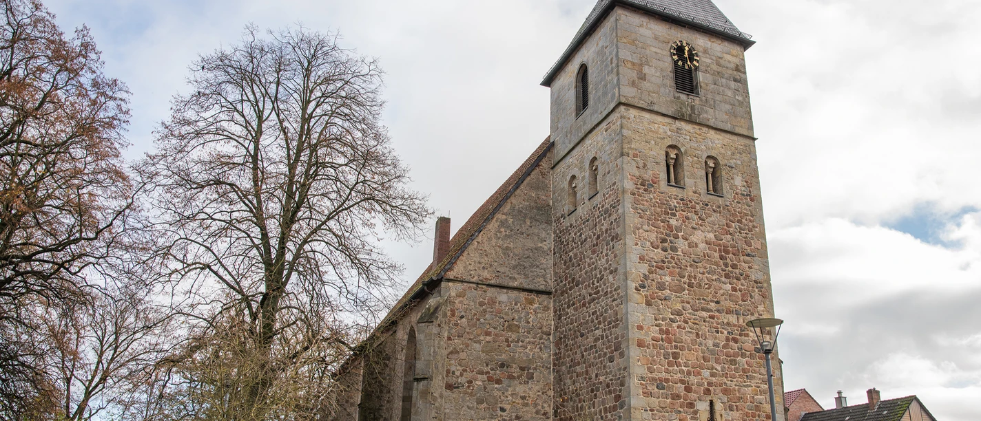 A historic sandstone church with a tower and a pointed roof, surrounded by trees and houses.