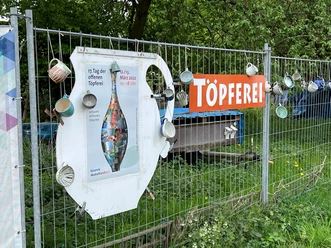 Ein Maschendrahtzaun mit bunten Tassen und einem Töpferplakat unter einem Töperei-Schild.