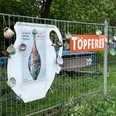 Töpferei Ein Maschendrahtzaun mit bunten Tassen und einem Töpferplakat unter einem Töperei-Schild.