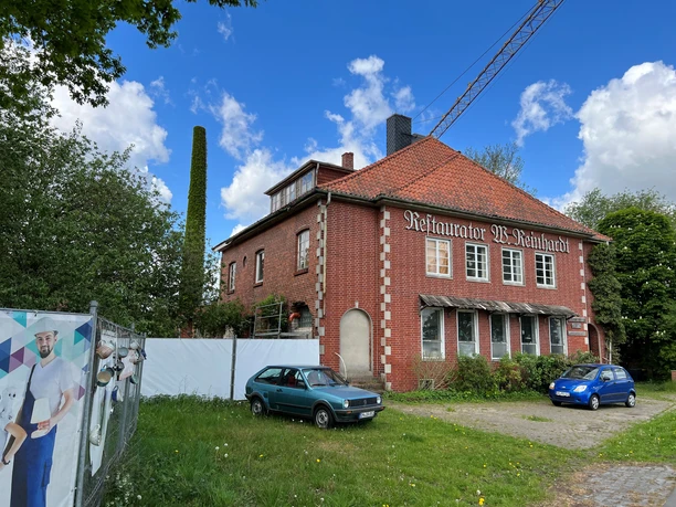 Breites Backsteingebäude mit rotem Ziegeldach, Fenstermarkisen und parkenden Autos, blauer Himmel.