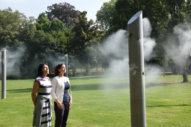 Menschen stehen in einem Park vor einer Nebelskulptur aus Metall, die Wassernebel sprüht.