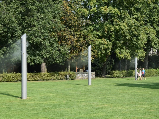 DSC_0174.JPG Dreiköpfige Familie spaziert auf grüner Wiese, umgeben von hohen, rauchenden Skulpturen und Bäumen.