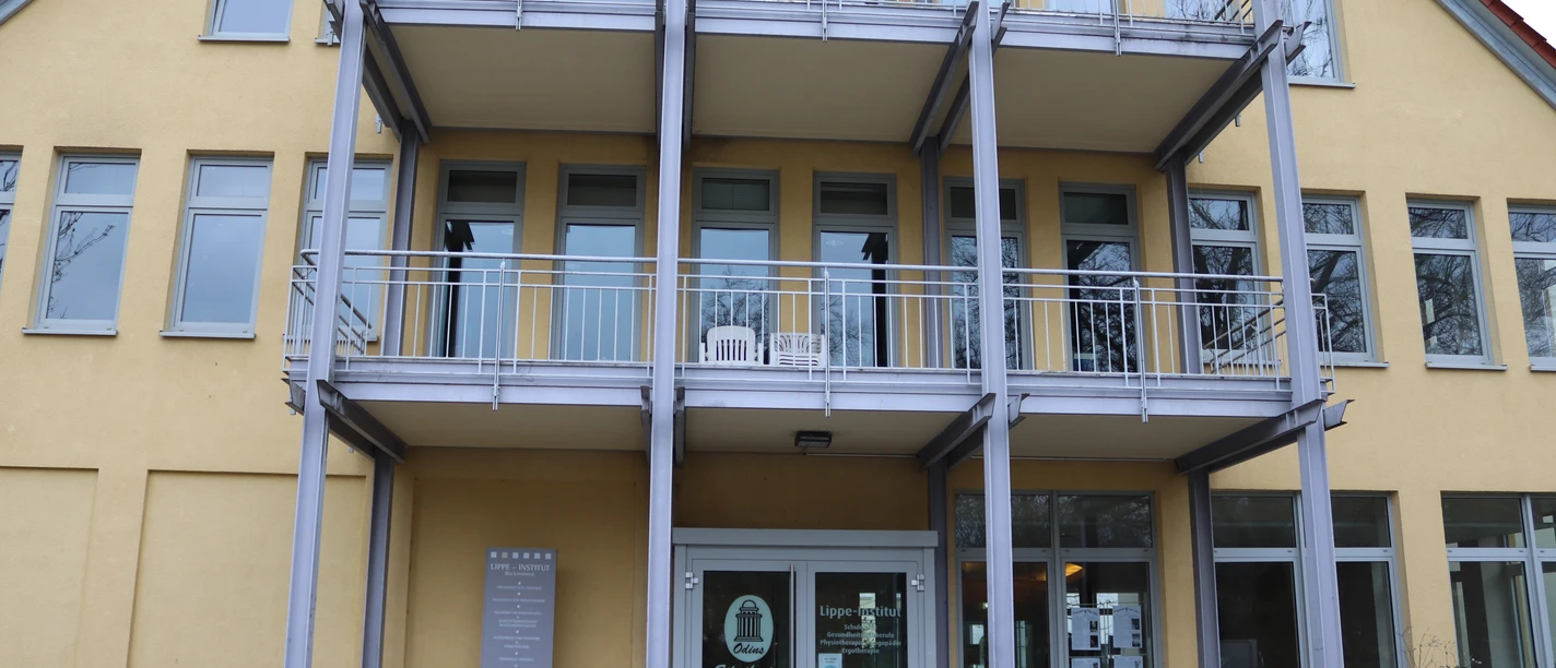 Blick auf den Eingang eines modernen, mehrstöckigen Gebäudes mit Balkonen und großen Fenstern.