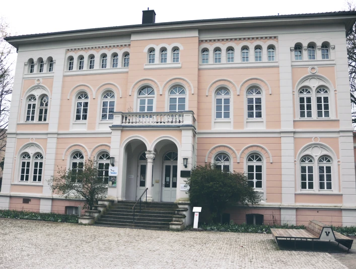 Ein historisches Gebäude mit rosafarbener Fassade, Bögen und vielen Fenstern auf Kopfsteinpflaster.