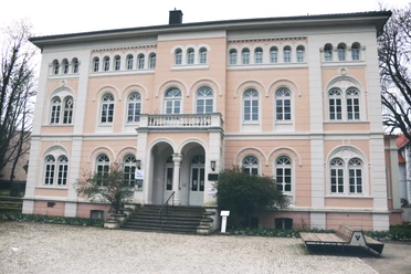 IMG_0420.JPG Ein historisches Gebäude mit rosafarbener Fassade, Bögen und vielen Fenstern auf Kopfsteinpflaster.
