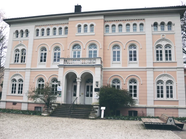 IMG_0420.JPG Ein historisches Gebäude mit rosafarbener Fassade, Bögen und vielen Fenstern auf Kopfsteinpflaster.