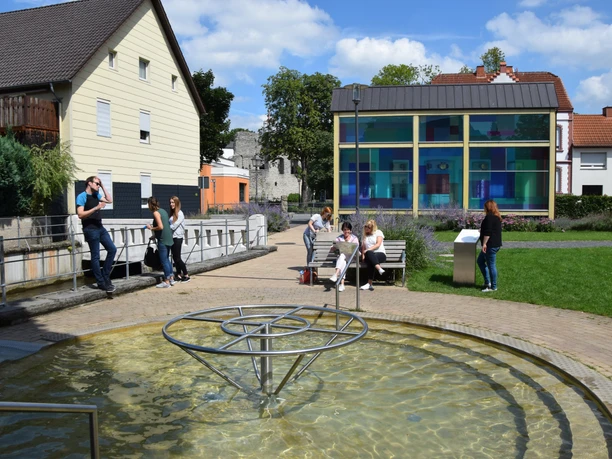 Personen entspannen in einer sonnigen Parkanlage mit Wasserspiel, moderner Glaskunst und Sitzgelegenheiten.