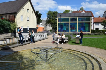 DSC_0288.JPG Personen entspannen in einer sonnigen Parkanlage mit Wasserspiel, moderner Glaskunst und Sitzgelegenheiten.
