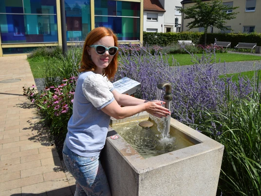 DSC_0298.JPG Eine Frau mit rotem Haar und Sonnenbrille wäscht ihre Hände an einem modernen Brunnen im Freien.