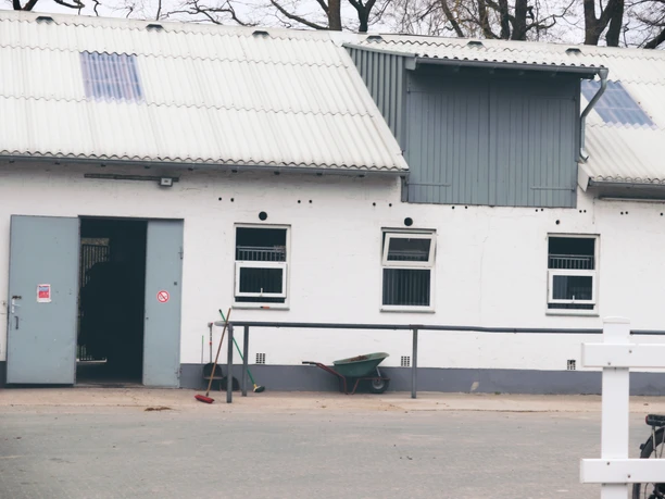 Weißes Gebäude mit Satteldach, mehrere kleine Fenster und offene Tür; Schubkarre und Besen davor.