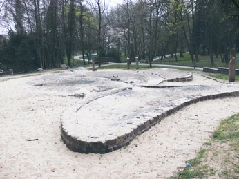 Ein weitläufiger Sandplatz im Park mit niedrigen gemauerten Strukturen unter kahlen Bäumen.