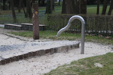 Wasserspielbereich mit schlangenförmigem Wasserhahn in einem Park, umgeben von Sand und Rasen.