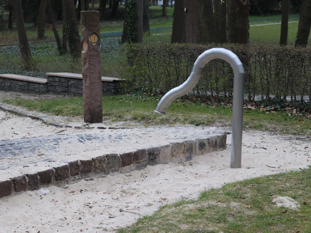 Wasserspielbereich mit schlangenförmigem Wasserhahn in einem Park, umgeben von Sand und Rasen.