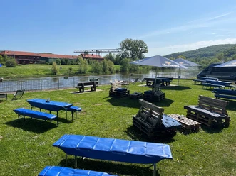Holzbänke und Tische aus Paletten stehen auf einer Wiese am Wasser. Ein blauer Himmel spannt sich darüber.