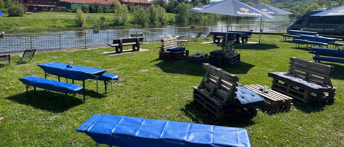 Sitzplätze Vanessas Hafenkante Holzbänke und Tische aus Paletten stehen auf einer Wiese am Wasser. Ein blauer Himmel spannt sich darüber.
