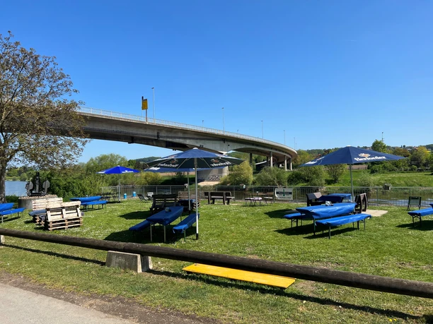 Picknickbereich am Ufer mit Biertischen, Sonnenschirmen und Blick auf eine Flussbrücke bei sonnigem Wetter.